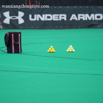 A sports field features a black and pink sports bag, two pyramids of yellow balls, and a portion of a person in dark athletic gear. The background displays a banner with the Under Armour logo.