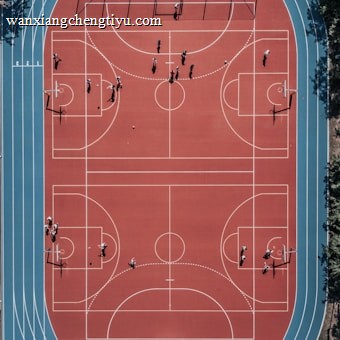 An aerial view of a large, vibrant sports complex featuring a red basketball court with multiple hoops and a surrounding blue running track. Several people are scattered across the area, engaged in various activities.