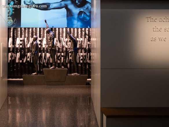A dimly lit display features statues of athletes standing on a podium. They are positioned in front of a backdrop with the words 'LEVELING THE PLAYING FIELD' in bold letters. On the wall to the right, a softly lit quote is partially visible. The overall ambiance suggests a museum or exhibition setting.