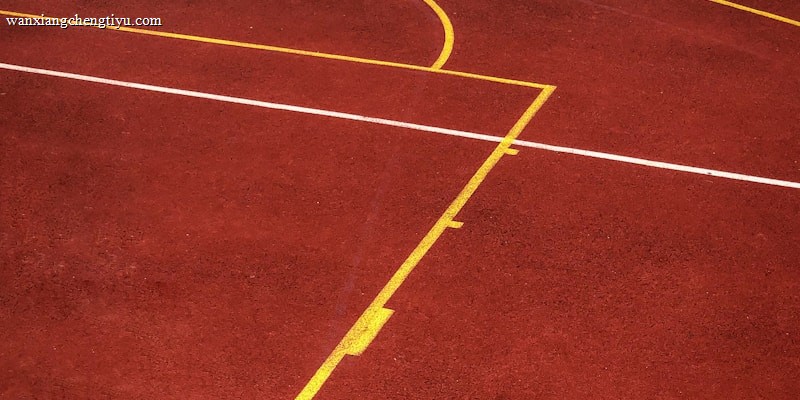 A sports court with a reddish-brown surface and bright yellow and white lines. The lines form geometric shapes, including rectangles and circles, suggesting markings for games like basketball or netball.