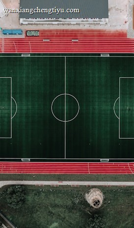 An aerial view of a large sports complex featuring a central green football field surrounded by a red running track. Adjacent to the track, there is a parking area, buildings, and pathways. Nearby, there are additional sports courts in different colors, including a blue swimming pool and orange basketball courts.