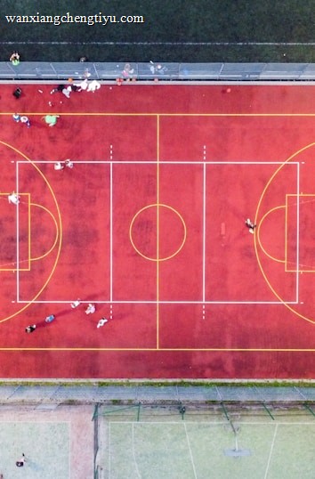 An aerial view of a sports court with a vibrant red surface and yellow lines marking different sections for basketball and soccer. Several people are present on the court, some engaged in activities, while others are standing along the perimeter. The court is enclosed by a metal fence and surrounded by grass on all sides.